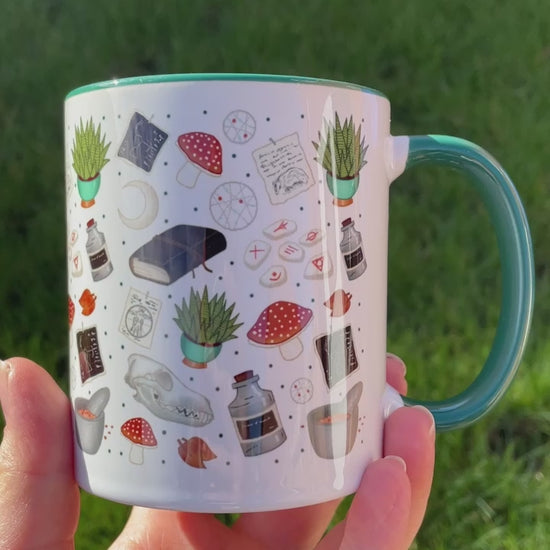 druid themed mug in green feature mushrooms, skulls, plants and runes in shades of greens and blues, being shown held in hand outside in the garden