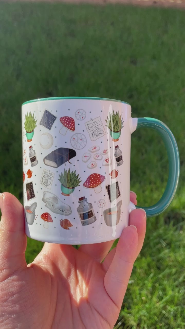 druid themed mug in green feature mushrooms, skulls, plants and runes in shades of greens and blues, being shown held in hand outside in the garden